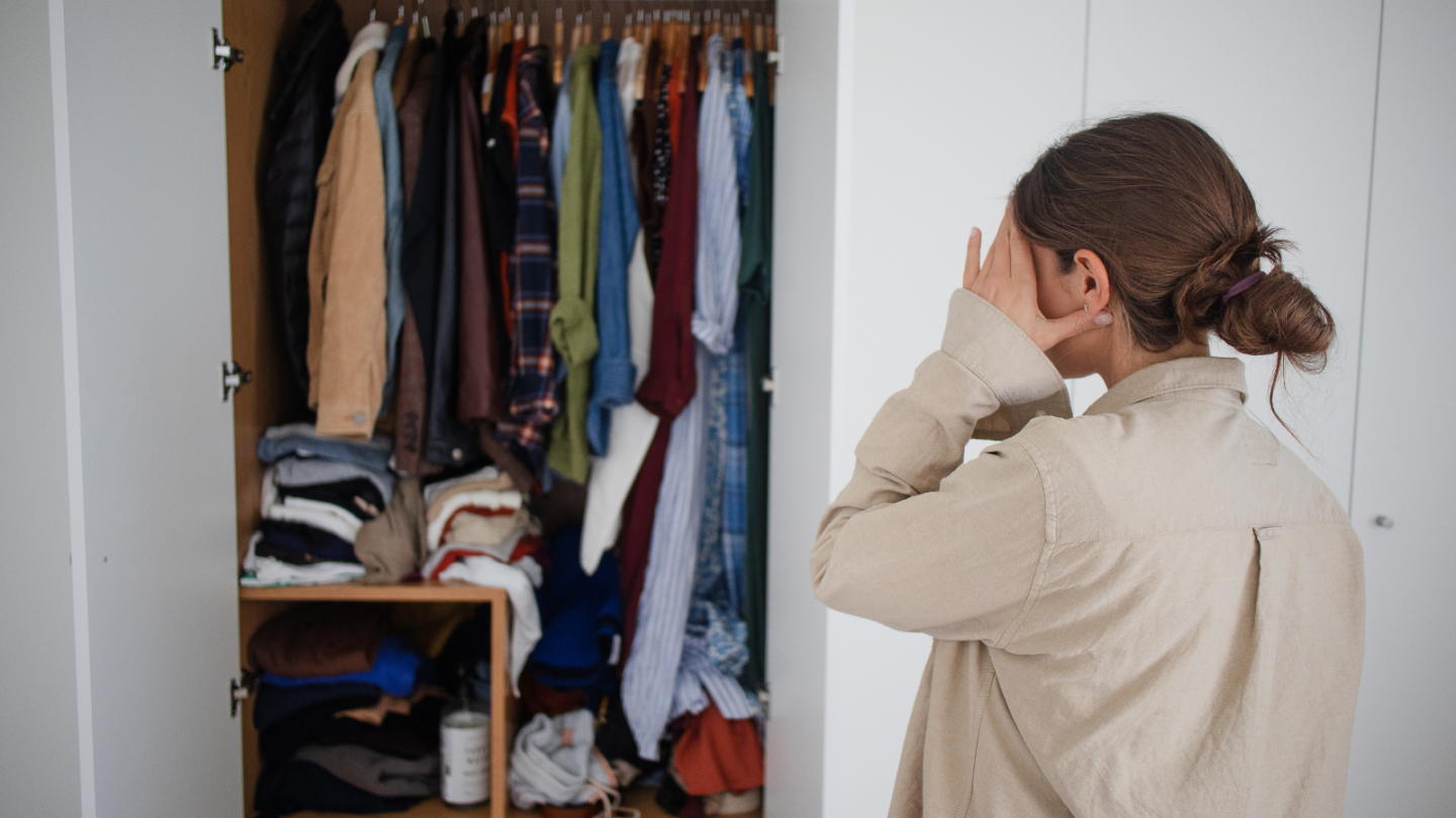 A lady with brown hair looking at her cluttered closet and feeling overwhelmed by the mess. She is thinking about the benefits of decluttering her closet will have on her mental health. 